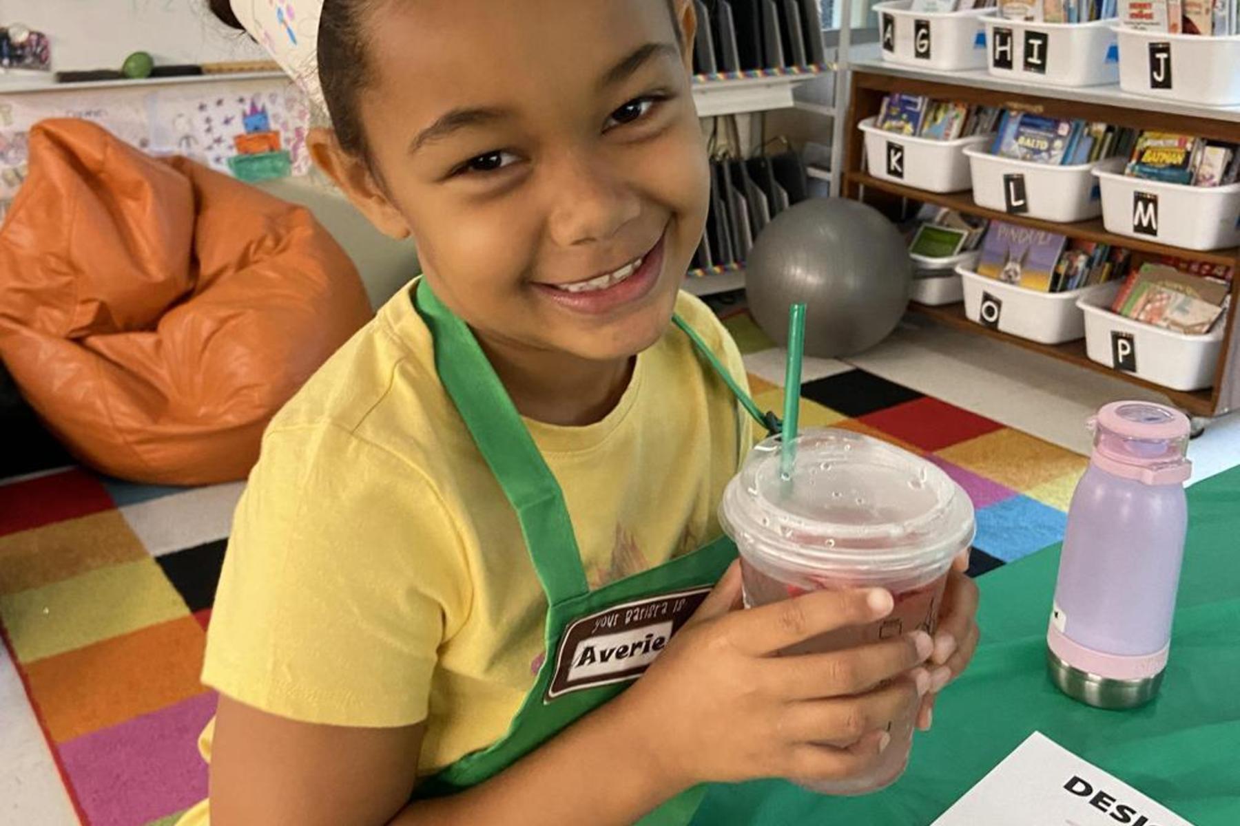 A smiling girl in a yellow shirt holding a drink, sitting at a colorful table.