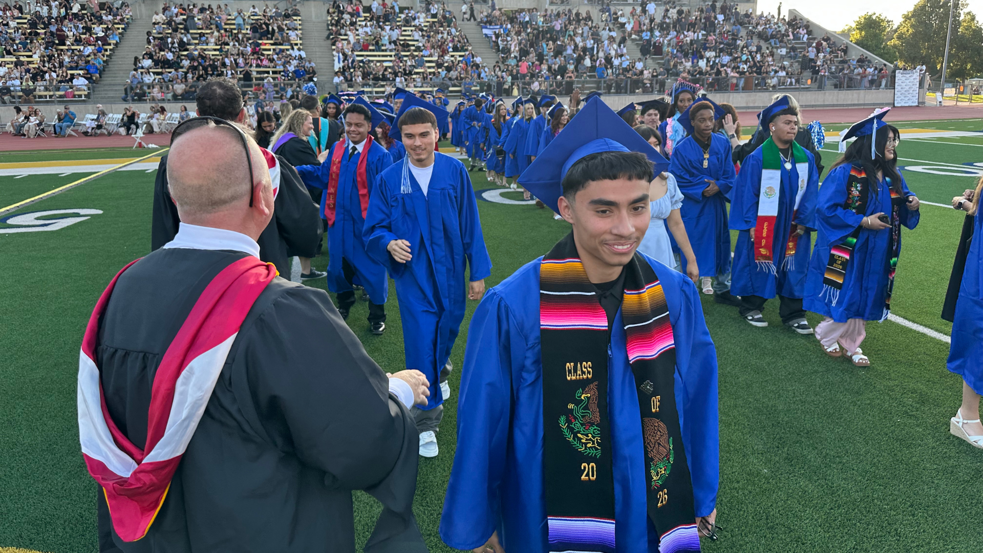 Graduates in blue gowns walking on a field during a ceremony.