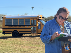 school staff member checking papers while a school bus pulls in behind them