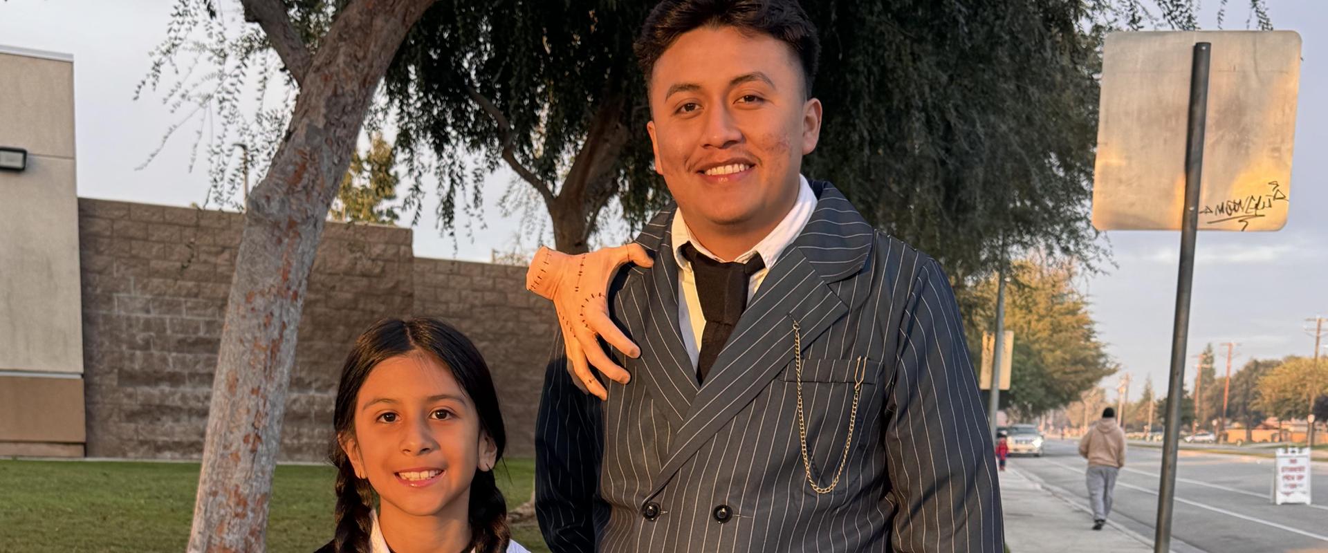 Man in pinstriped suit stands with girl in black dress outdoors at sunset.